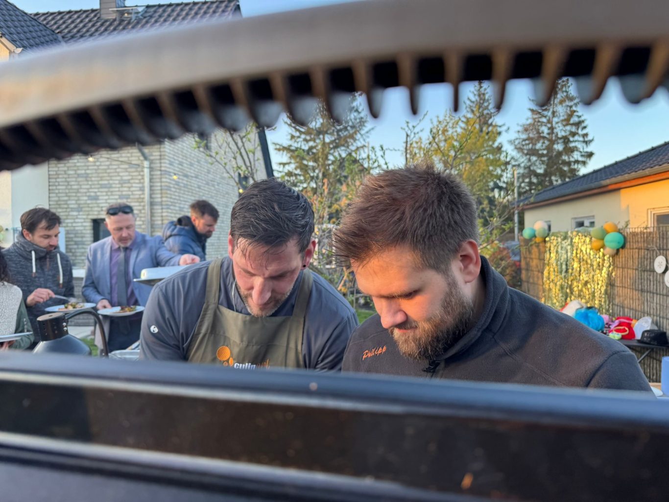 Zwei Köche von CulinART arbeiten am Grill, im Hintergrund Gäste mit Tellern bei einer Gartenfeier