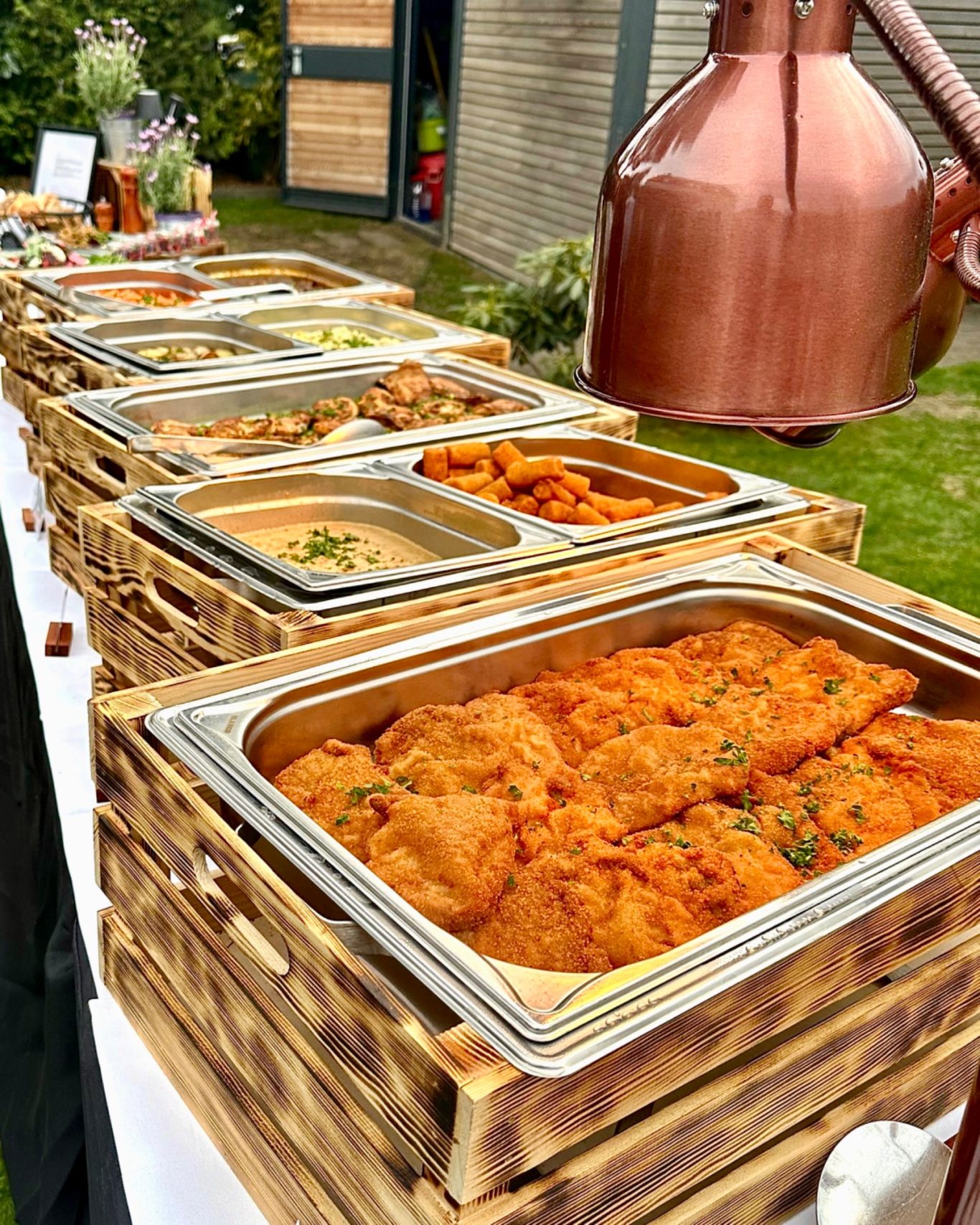 Warmes Buffet im Garten: Schnitzel, Kroketten und Beilagen in Edelstahlbehältern auf geflammten Holzkisten